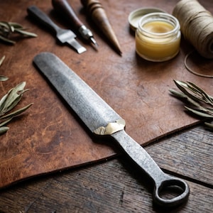 May include: A hand-forged steel leatherworking creaser tool featuring a long, flat, textured blade with a polished brass bolster and a circular ring handle, designed for manual leather crafting and edge finishing tasks.