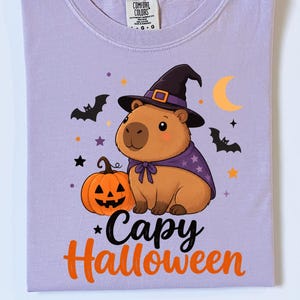 Puede incluir: Una camiseta de color morado claro con un gr&aacute;fico de un capibara marr&oacute;n con un sombrero de bruja negro y una capa morada. Junto al capibara hay una calabaza naranja, murci&eacute;lagos negros, estrellas y una luna creciente amarilla. Texto: Capy Halloween.