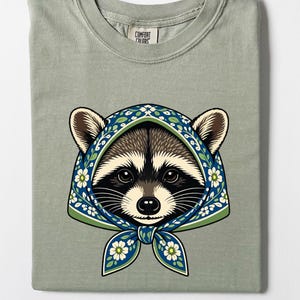 Könnte beinhalten: Ein salbeigrünes T-Shirt mit einem Grafikdruck eines Waschbären, der ein blau-grünes florales Kopftuch trägt, das unter dem Kinn gebunden ist. Das T-Shirt hat einen Rundhalsausschnitt und ein Comfort Colors Markenetikett im Kragenbereich.