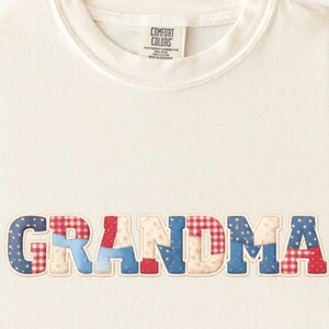 May include: Cream-colored t-shirt with the word "GRANDMA" appliquéd across the front. The letters are made of patchwork fabric in red, white, and blue patterns. The shirt has a crew neck and a tag that says "COMFORT COLORS".