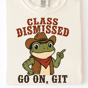 Może przedstawiać: Kremowy t-shirt z grafiką zielonej żaby w brązowym kowbojskim kapeluszu, kamizelce i czerwonej bandanie. Napis nad żabą brzmi "CLASS DISMISSED", a napis poniżej to "GO ON, GIT" wykonany ciemnobrązową czcionką o postarzanym wyglądzie.