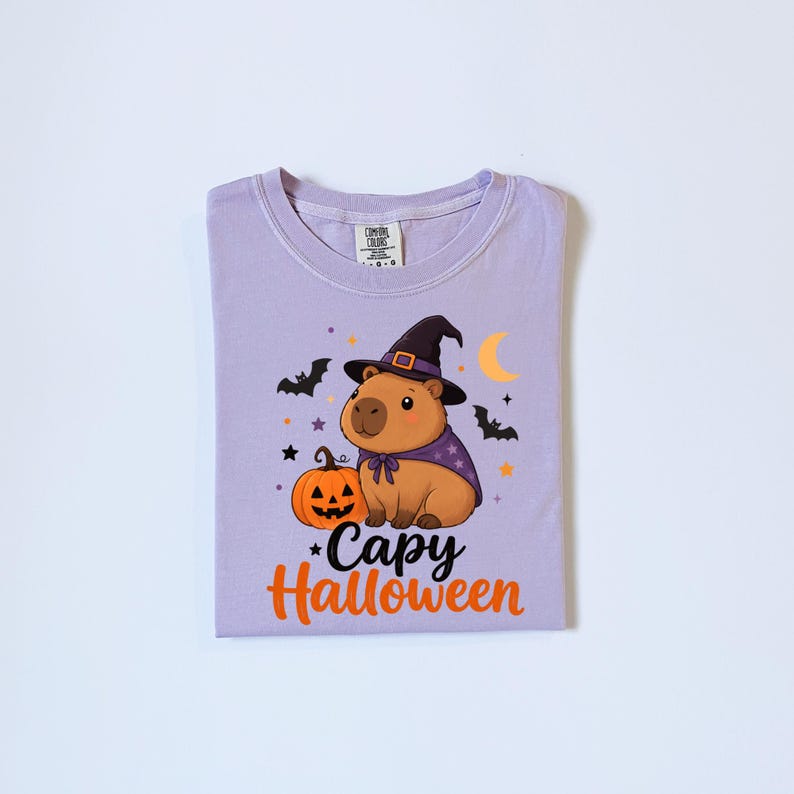 Puede incluir: Una camiseta de color morado claro con un gr&aacute;fico de un capibara marr&oacute;n con un sombrero de bruja negro y una capa morada. Junto al capibara hay una calabaza naranja, murci&eacute;lagos negros, estrellas y una luna creciente amarilla. Texto: Capy Halloween.