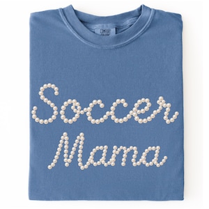 May include: A blue t-shirt with the words "Soccer Mama" spelled out in white pearl-like beads. The shirt is folded neatly, and the brand "Comfort Colors" is visible on the tag.