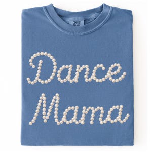 May include: A blue t-shirt with the words "Dance Mama" spelled out in white pearl-like beads. The shirt has a crew neck and is folded neatly. The brand "Comfort Colors" is visible on the tag.