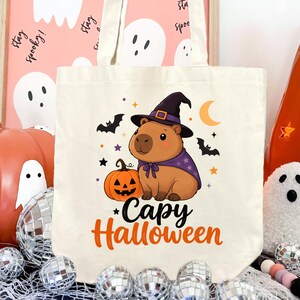 Puede incluir: Una bolsa de tela color crema con el gr&aacute;fico de un capibara marr&oacute;n que lleva un sombrero de bruja y una capa morada, sentado junto a una calabaza. El dise&ntilde;o incluye murci&eacute;lagos, estrellas, una luna creciente y el texto Capy Halloween en naranja.