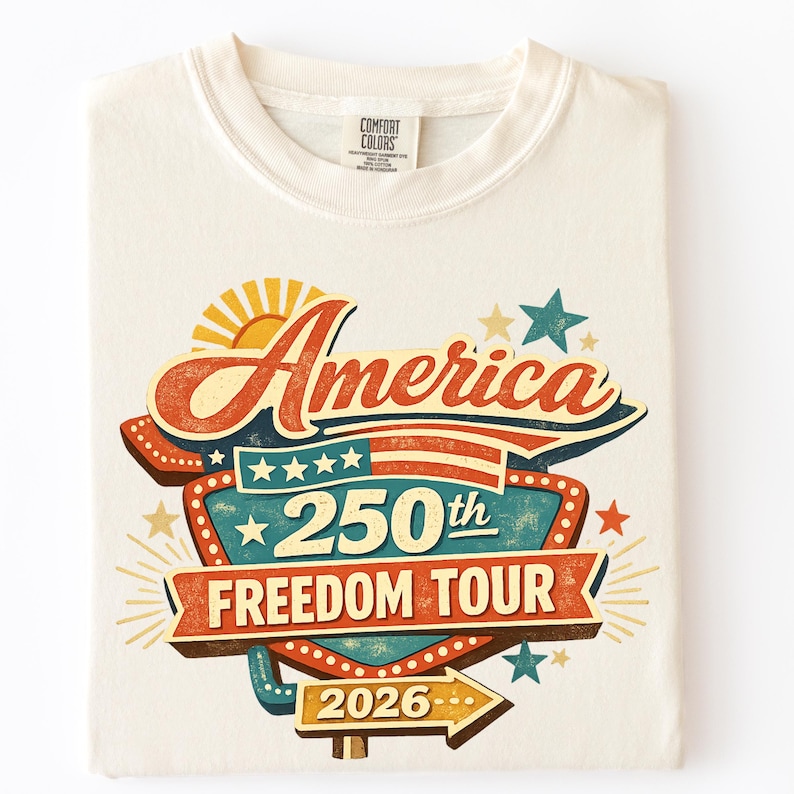 May include: Cream-colored t-shirt with a retro design celebrating America's 250th anniversary. The graphic includes the word "America" in a vintage font, a sun, stars, and the text "250th Freedom Tour 2026."