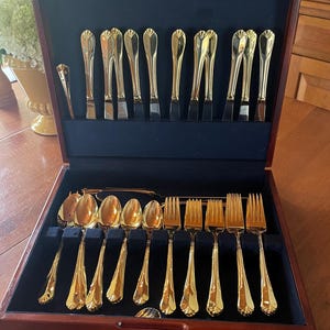 May include: A gold-toned flatware set arranged in a dark wood storage case with a navy blue velvet interior. The set includes knives, spoons, and forks, each featuring a smooth, reflective finish and a simple, tapered handle design.