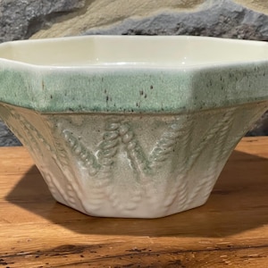 May include: An octagonal ceramic bowl featuring a cream-coloured interior and a light green exterior glaze. The exterior surface displays a textured, raised rope-like pattern around the sides, with a speckled finish near the top rim.
