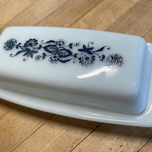 May include: A rectangular white milk glass butter dish with a lid. The lid features a dark blue floral pattern running horizontally across the center. The dish has a smooth, glossy finish and a simple, clean design with rounded edges on the base.
