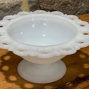 Puede incluir: Un cuenco blanco con pedestal de estilo vintage. El cuenco tiene un borde festoneado con una serie de círculos abiertos. El cuenco se asienta sobre una base redonda. El cuenco está hecho de vidrio opaco.