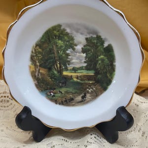 May include: White porcelain dish with a scalloped edge and gold trim. The center features a pastoral landscape painting with trees, figures, and animals. The dish is displayed on a black stand.