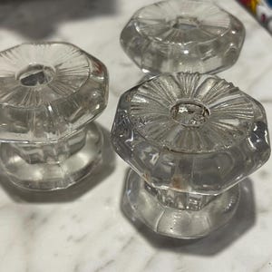 May include: Three clear glass cabinet knobs featuring an octagonal shape with a faceted, sunburst-like pattern radiating from the center. Each knob has a smooth, rounded base and a central circular indentation, showcasing a transparent, reflective finish.