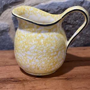 May include: A yellow and white speckled ceramic pitcher with a black rim and handle. The pitcher has a curved spout and a rounded body. The handle is black with a white interior. The pitcher is sitting on a wooden surface.