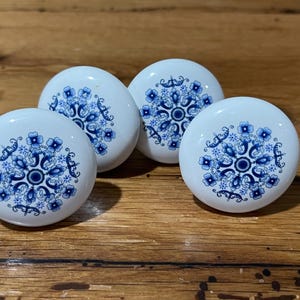 May include: Four white ceramic cabinet knobs with a blue floral design. The knobs are round and have a glossy finish. The blue pattern is symmetrical and detailed, with a central circle and surrounding floral elements.
