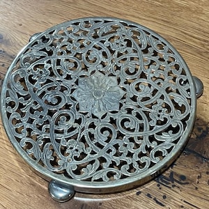 Op de afbeelding: Een zilverkleurige, ronde onderzetter met een ingewikkeld opengewerkt ontwerp. De onderzetter heeft een centraal bloemmotief en decoratief krulwerk. Het heeft drie kleine, afgeronde voetjes voor verhoging en is ontworpen om oppervlakken tegen hitte te beschermen.