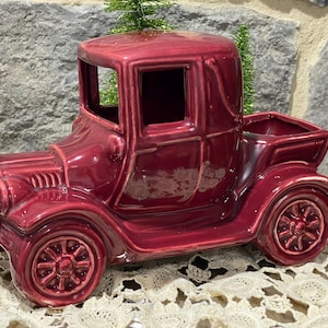 May include: A glossy, deep red ceramic vintage truck figurine featuring a hollowed-out truck bed, detailed spoked wheels, a front grille, and a cabin with open windows. The truck has a smooth, reflective finish and a classic, antique vehicle design.