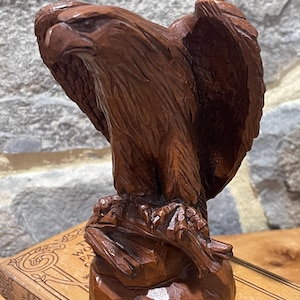May include: A hand-carved wooden eagle sculpture with outstretched wings. The eagle is perched on a rough-hewn base, showcasing detailed feather textures and a focused gaze. The sculpture is a rich brown color, suggesting a natural wood finish.