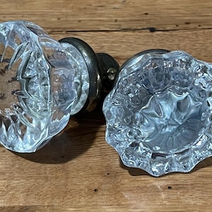 May include: Two clear, faceted glass door knobs with brass hardware. The knobs have a rounded, floral-like design. The knobs are displayed on a wooden surface.