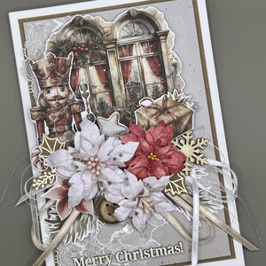 以下が含まれることがあります： 手作りのクリスマスカード。くるみ割り人形、赤いカーテンのある窓、ギフトボックスの詳細なイラストが特徴です。白と赤のポインセチア、木の雪の結晶、そして「Merry Christmas！」の文字で飾られています。