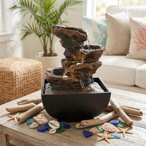 May include: An indoor water feature with a layered, rock-effect design. The fountain is dark brown and set in a black bowl. Driftwood, seashells, and sea glass are arranged around the base, creating a coastal theme.
