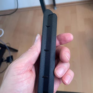 May include: A matte black, elongated, rectangular plastic component with rounded ends. The object features a raised central ridge running along its length, punctuated by three small, evenly spaced perpendicular fins or tabs protruding from the side.