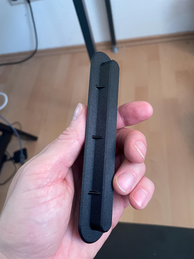 May include: A matte black, elongated, rectangular plastic component with rounded ends. The object features a raised central ridge running along its length, punctuated by three small, evenly spaced perpendicular fins or tabs protruding from the side.