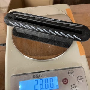 May include: A black, elongated, rectangular object with a central slot and a textured, diagonal-striped pattern on its top surface, resting on a digital scale that displays a weight of 28.00 grams. The object features a small circular hole at one end.