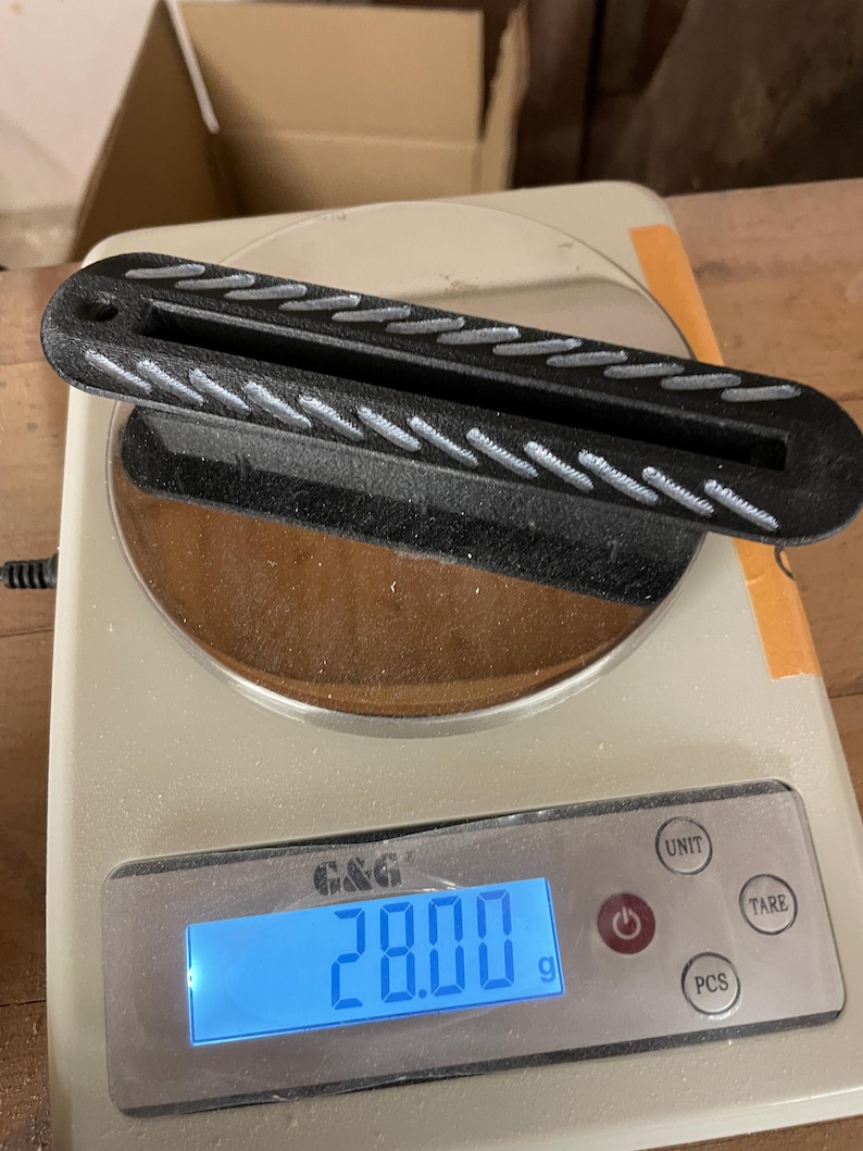 May include: A black, elongated, rectangular object with a central slot and a textured, diagonal-striped pattern on its top surface, resting on a digital scale that displays a weight of 28.00 grams. The object features a small circular hole at one end.