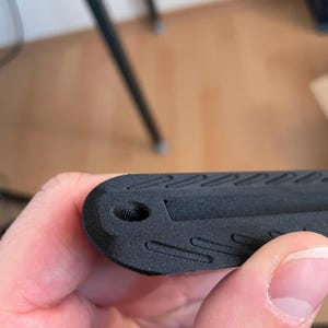 May include: A matte black 3D printed object with a rounded end featuring a small circular hole and a series of recessed diagonal grooves along the surface. The texture shows visible layer lines consistent with additive manufacturing processes.