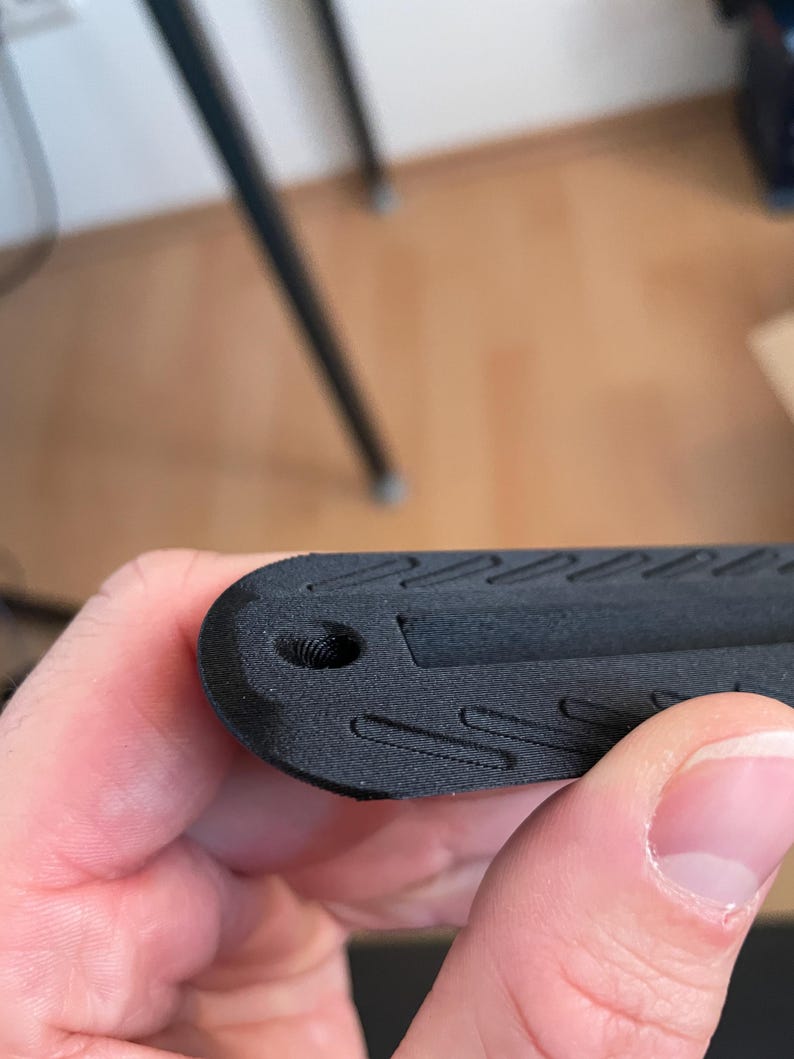 May include: A matte black 3D printed object with a rounded end featuring a small circular hole and a series of recessed diagonal grooves along the surface. The texture shows visible layer lines consistent with additive manufacturing processes.