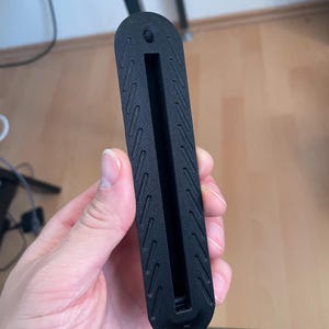May include: A matte black, elongated, rectangular component with rounded ends, featuring a central vertical slot and a small threaded hole at the top. The surface is textured with a repeating pattern of diagonal, parallel, recessed lines.