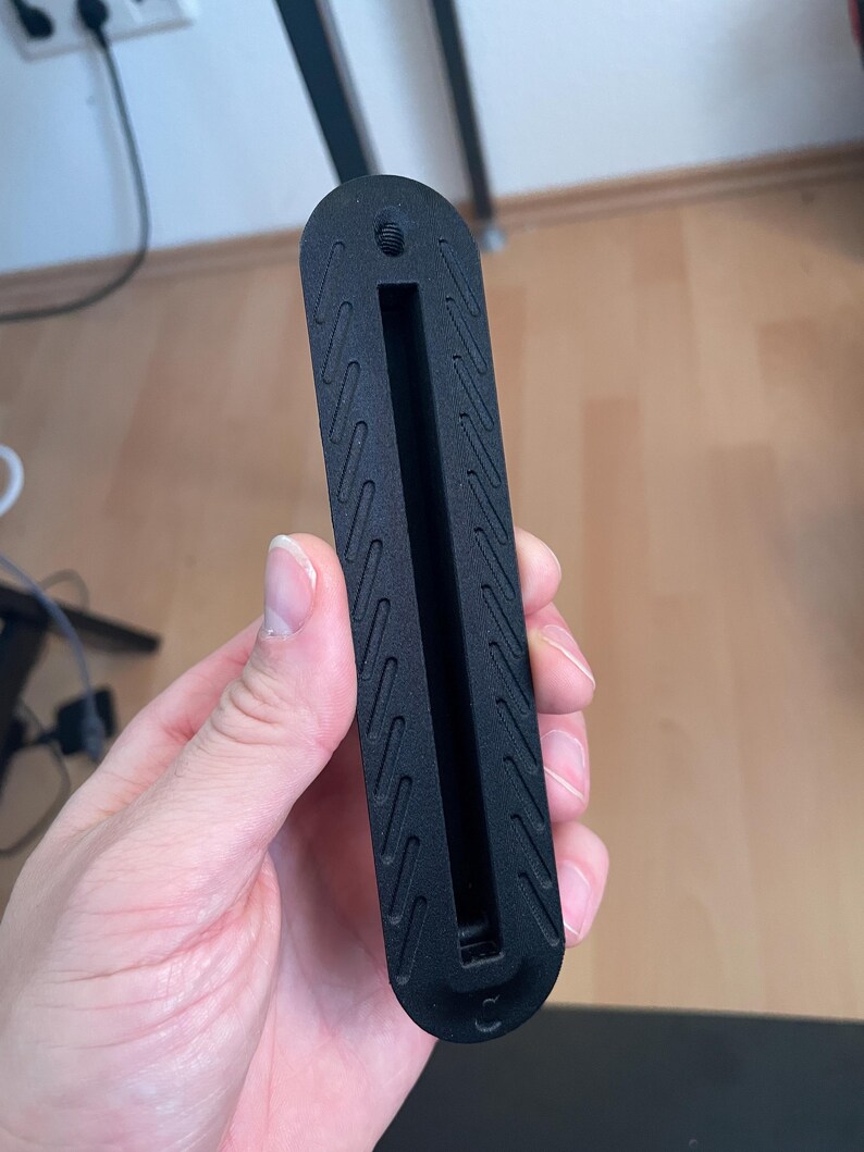 May include: A matte black, elongated, rectangular component with rounded ends, featuring a central vertical slot and a small threaded hole at the top. The surface is textured with a repeating pattern of diagonal, parallel, recessed lines.