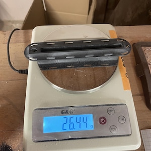 May include: A black, rectangular plastic component with a central slot and perforated edges sits on a white digital scale. The scale display shows a weight of 26.44 grams. The device features buttons labeled UNIT, TARE, and PCS, with a blue backlight.