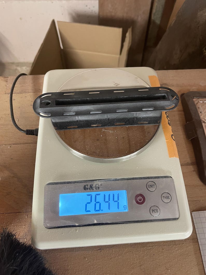 May include: A black, rectangular plastic component with a central slot and perforated edges sits on a white digital scale. The scale display shows a weight of 26.44 grams. The device features buttons labeled UNIT, TARE, and PCS, with a blue backlight.