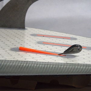 May include: A close-up of a surfboard with a gray and white honeycomb pattern. The board has an orange fin key and a black fin. The board has a gray edge and a white background.