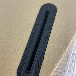 May include: A black plastic surfboard fin box embedded into a tan foam surfboard surface. The fin box features a long, narrow central slot for fin attachment and a textured border with a repeating diagonal pattern and a single small metal screw.
