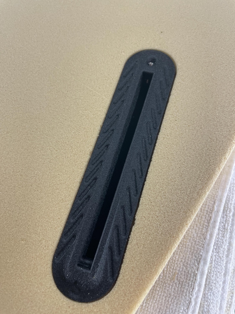 May include: A black plastic surfboard fin box embedded into a tan foam surfboard surface. The fin box features a long, narrow central slot for fin attachment and a textured border with a repeating diagonal pattern and a single small metal screw.
