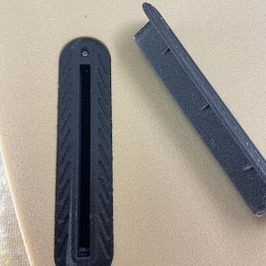 May include: A tan surfboard surface featuring a black plastic fin box with a textured border and a single screw hole. Beside the installed fin box lies a matching black plastic fin box filler piece with a rectangular shape and textured edges.