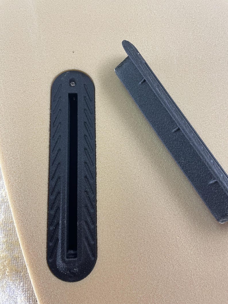 May include: A tan surfboard surface featuring a black plastic fin box with a textured border and a single screw hole. Beside the installed fin box lies a matching black plastic fin box filler piece with a rectangular shape and textured edges.