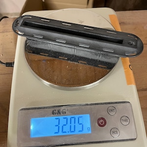 May include: A black, elongated, rectangular plastic component with a central slot and rounded ends, featuring a series of small rectangular cutouts along its perimeter, resting on a digital scale displaying a weight of 32.05 grams.