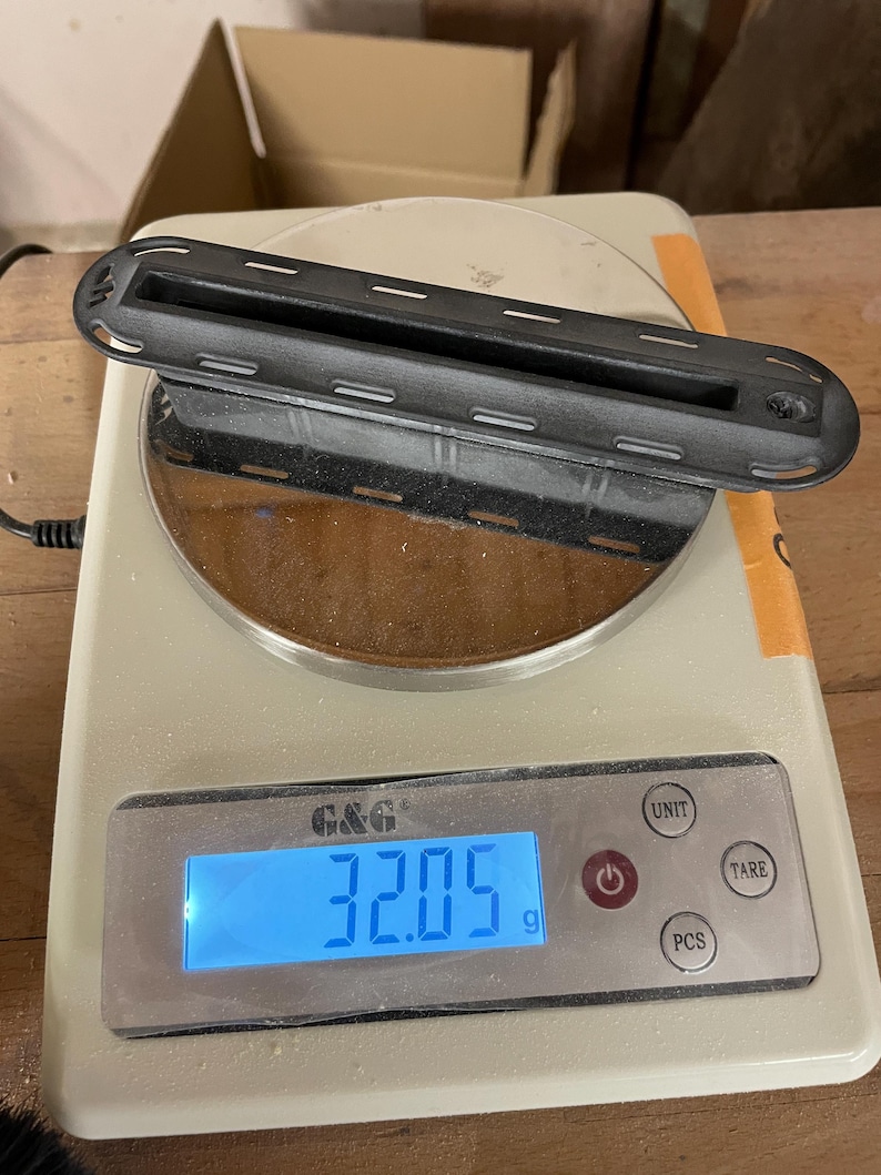 May include: A black, elongated, rectangular plastic component with a central slot and rounded ends, featuring a series of small rectangular cutouts along its perimeter, resting on a digital scale displaying a weight of 32.05 grams.
