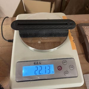May include: A black, elongated, textured plastic object with a central slot and a small circular hole at one end, resting on a white digital scale that displays a weight of 22.13 grams on its blue backlit screen, with the brand name G&G visible.