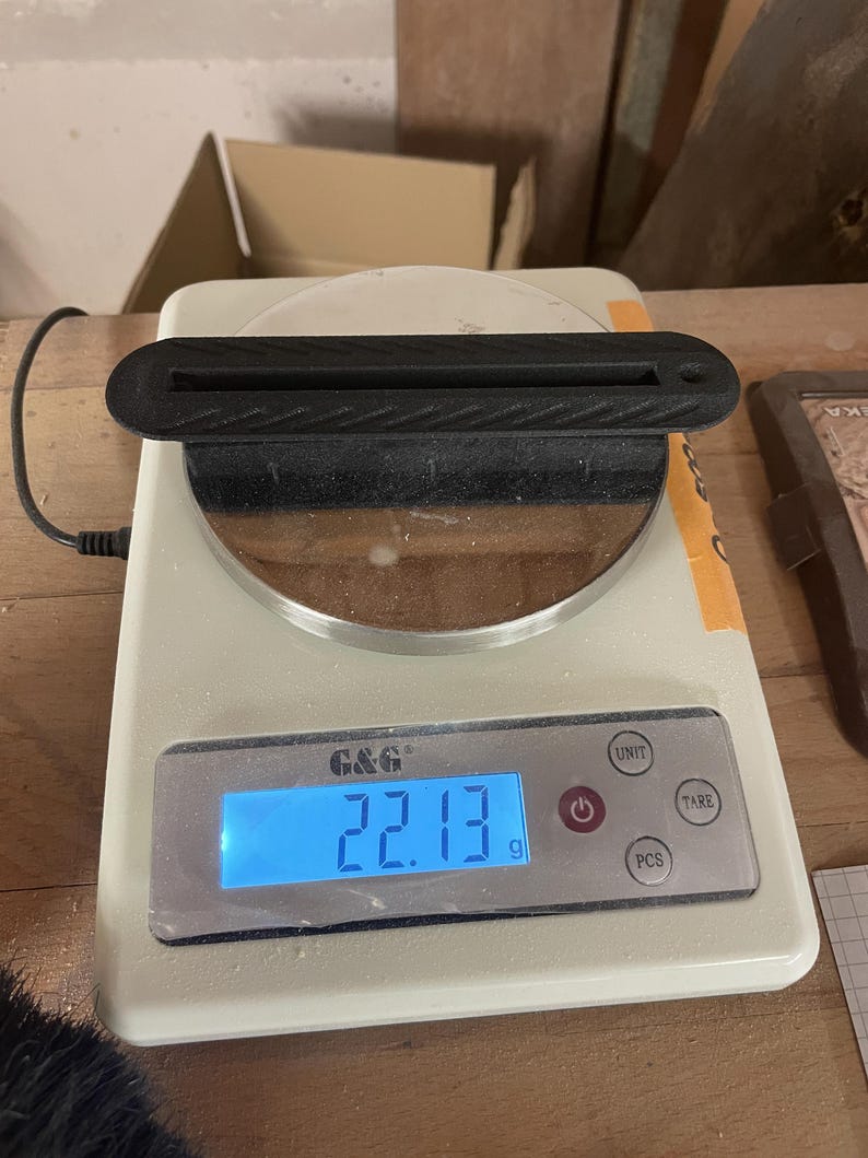 May include: A black, elongated, textured plastic object with a central slot and a small circular hole at one end, resting on a white digital scale that displays a weight of 22.13 grams on its blue backlit screen, with the brand name G&G visible.