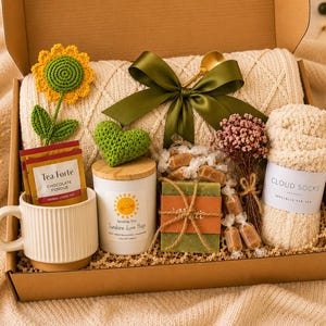 Könnte beinhalten: Eine Geschenkbox mit einer cremefarbenen Decke, einer Tasse mit Tee, einer Kerze, einem Paar Wolkensocken und einem Stück Seife. Die Box enthält auch eine grüne Schleife, eine gehäkelte Sonnenblume und ein gehäkeltes Herz.