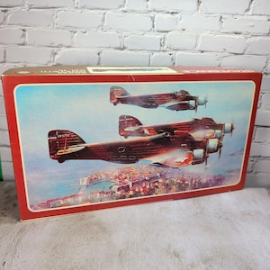 May include: A rectangular cardboard model kit box featuring a painting of three brown military aircraft flying in formation over a coastal city. The box has a red border and displays the text SAVOIA MARCHETTI SM 79 on the top edge.