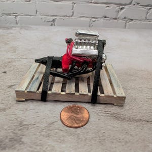 May include: A miniature model of a V8 engine with a chrome supercharger, red engine block, and black exhaust headers, secured with black straps to a small wooden pallet. A copper-colored United States penny is placed in front for scale.