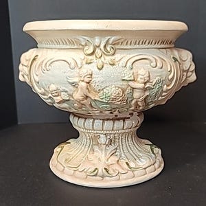 May include: A cream-colored ceramic pedestal bowl featuring a relief design of cherubs and foliage. The surface has a crackled glaze finish with subtle green and gold accents. The bowl has a wide rim, a flared body, and a sturdy, ornate base.