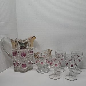 Op de afbeelding: Een heldere glazen kan, melkkan en vier wijnglazen. De set heeft een decoratief patroon met roze accenten en gouden rand. De kan heeft een handvat en een uitlopende rand. De glazen hebben een voet.