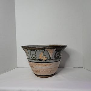 May include: A handcrafted ceramic bowl with a wide, flared rim. The bowl features a textured, light brown base, a gray band with leaf-like designs, and a dark brown rim. The design is symmetrical and the bowl is likely for decorative use.
