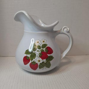 Könnte beinhalten: Ein weißer Keramikkrug mit einem dekorativen Muster aus roten Erdbeeren, grünen Blättern und kleinen weißen Blüten. Der Krug verfügt über einen geschwungenen Henkel mit einem Schnörkel am oberen Ende und einen breiten, ausgestellten Ausguss.